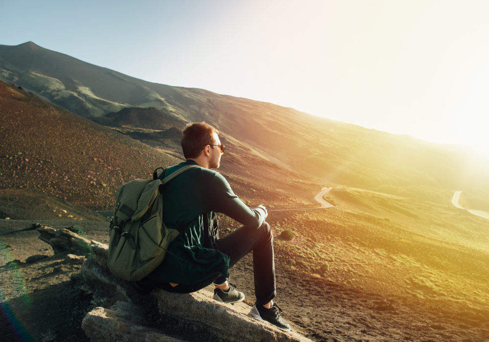 Man with rucksack sitting on rock at sunset on background of volcano Etna mountain in Sicily